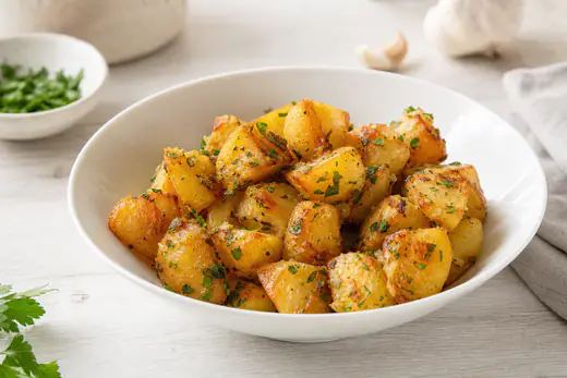 Kullanruskeat valkosipuliperunapalat persiljan ja valkosipulivoin kera valkoisessa kulhossa